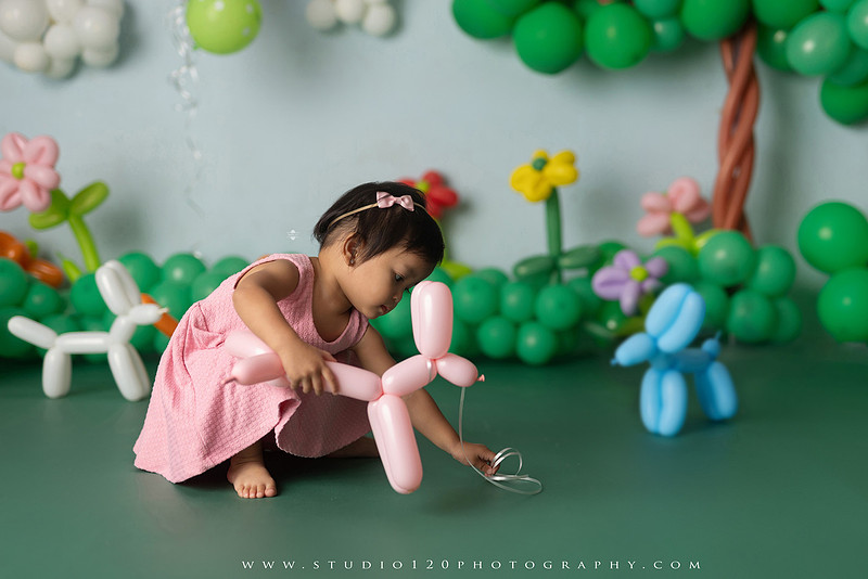 Art-inspired two-year-old birthday photo session featuring a toddler in a pink dress within a whimsical balloon garden set, with playful sculptural elements and a visual nod to Jeff Koons