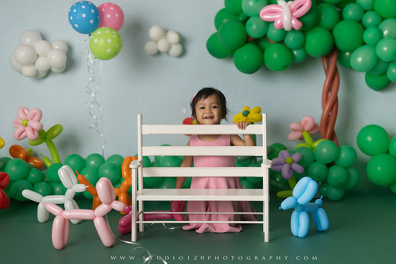 Art-inspired two-year-old birthday photo session featuring a toddler in a pink dress within a whimsical balloon garden set, with playful sculptural elements and a visual nod to Jeff Koons