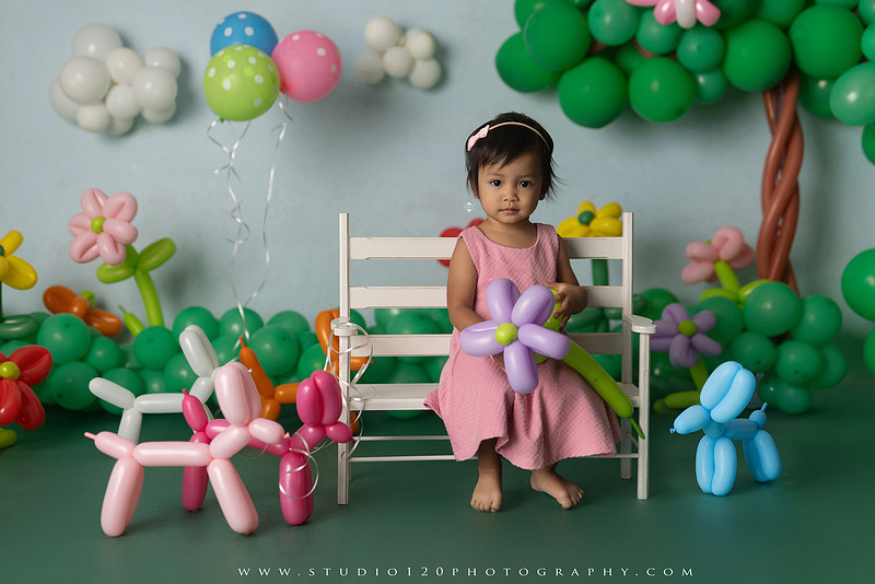 Art-inspired two-year-old birthday photo session featuring a toddler in a pink dress within a whimsical balloon garden set, with playful sculptural elements and a visual nod to Jeff Koons