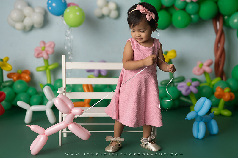 Art-inspired two-year-old birthday photo session featuring a toddler in a pink dress within a whimsical balloon garden set, with playful sculptural elements and a visual nod to Jeff Koons