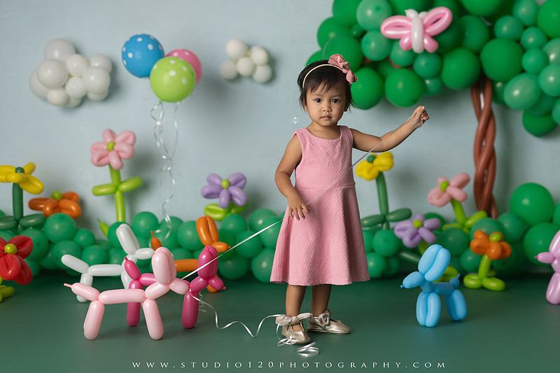 Art-inspired two-year-old birthday photo session featuring a toddler in a pink dress within a whimsical balloon garden set, with playful sculptural elements and a visual nod to Jeff Koons