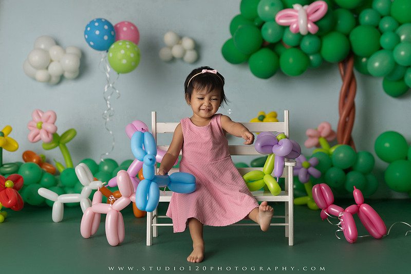 Art-inspired two-year-old birthday photo session featuring a toddler in a pink dress within a whimsical balloon garden set, with playful sculptural elements and a visual nod to Jeff Koons