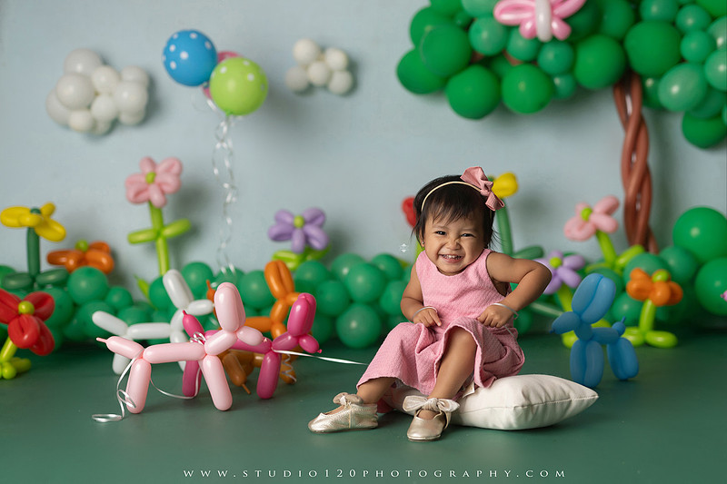 Art-inspired two-year-old birthday photo session featuring a toddler in a pink dress within a whimsical balloon garden set, with playful sculptural elements and a visual nod to Jeff Koons