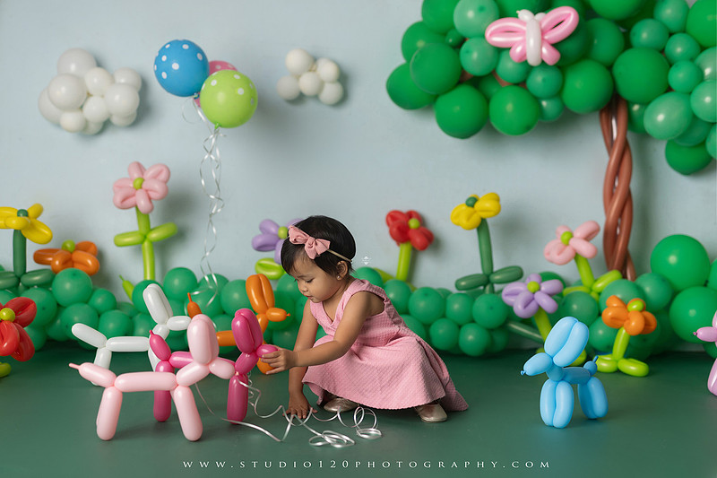 Art-inspired two-year-old birthday photo session featuring a toddler in a pink dress within a whimsical balloon garden set, with playful sculptural elements and a visual nod to Jeff Koons