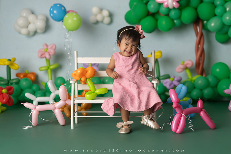 Art-inspired two-year-old birthday photo session featuring a toddler in a pink dress within a whimsical balloon garden set, with playful sculptural elements and a visual nod to Jeff Koons
