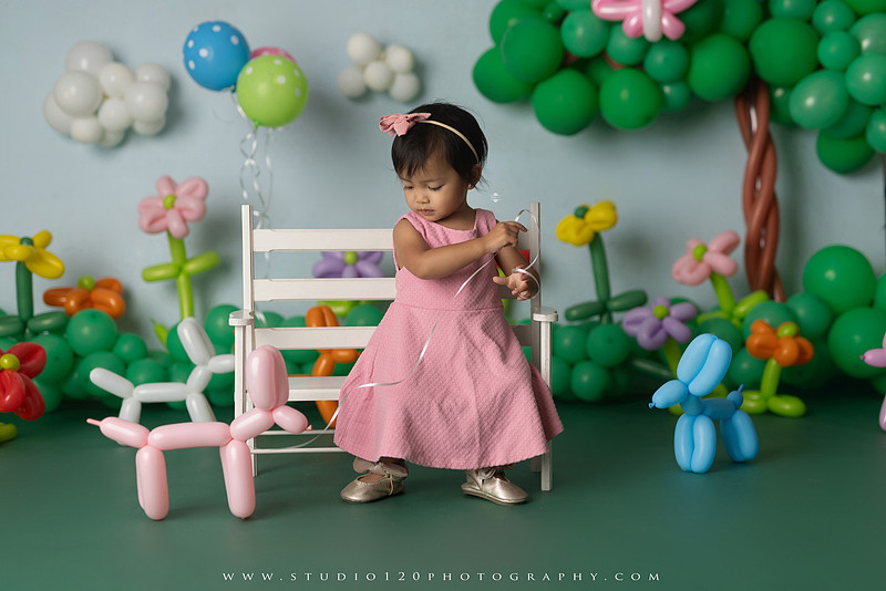Art-inspired two-year-old birthday photo session featuring a toddler in a pink dress within a whimsical balloon garden set, with playful sculptural elements and a visual nod to Jeff Koons