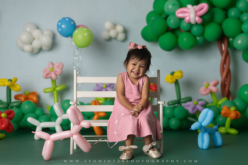 Art-inspired two-year-old birthday photo session featuring a toddler in a pink dress within a whimsical balloon garden set, with playful sculptural elements and a visual nod to Jeff Koons