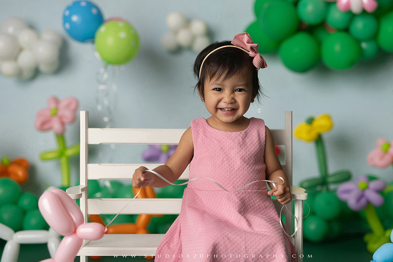 Art-inspired two-year-old birthday photo session featuring a toddler in a pink dress within a whimsical balloon garden set, with playful sculptural elements and a visual nod to Jeff Koons