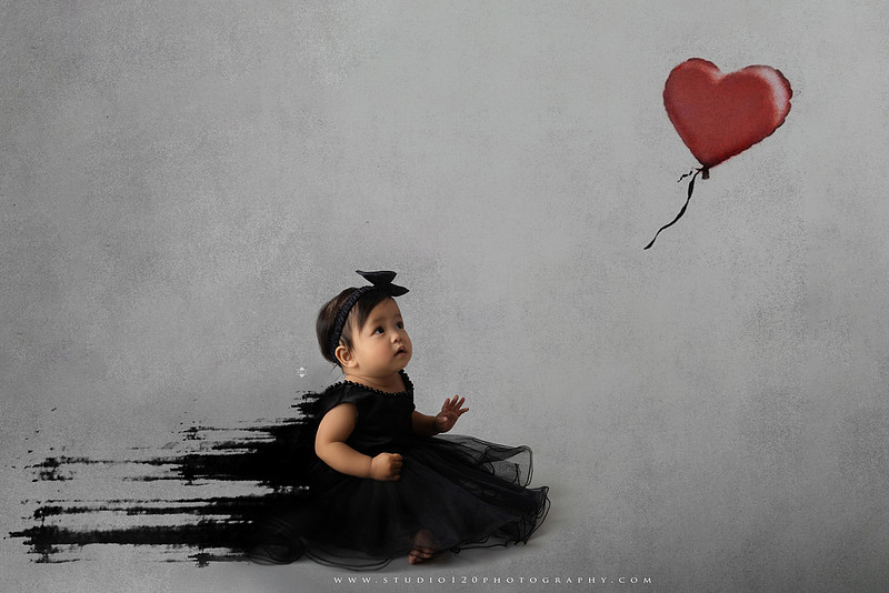 Banksy-inspired newborn photography featuring fine art minimalist styling with angel wings, painted motion effects, and a red heart balloon on a soft gray backdrop