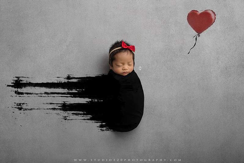 Banksy-inspired newborn photography featuring fine art minimalist styling with angel wings, painted motion effects, and a red heart balloon on a soft gray backdrop