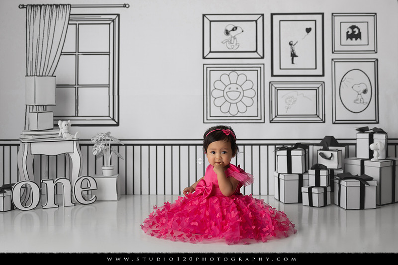 Art-inspired first birthday photo session featuring a toddler in a vibrant pink dress against a graphic black-and-white illustrated set, with visual nods to Takashi Murakami, KAWS, Jeff Koons, and Invader
