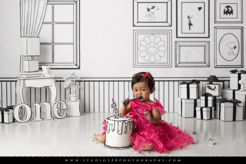 Art-inspired first birthday photo session featuring a toddler in a vibrant pink dress against a graphic black-and-white illustrated set, with visual nods to Takashi Murakami, KAWS, Jeff Koons, and Invader
