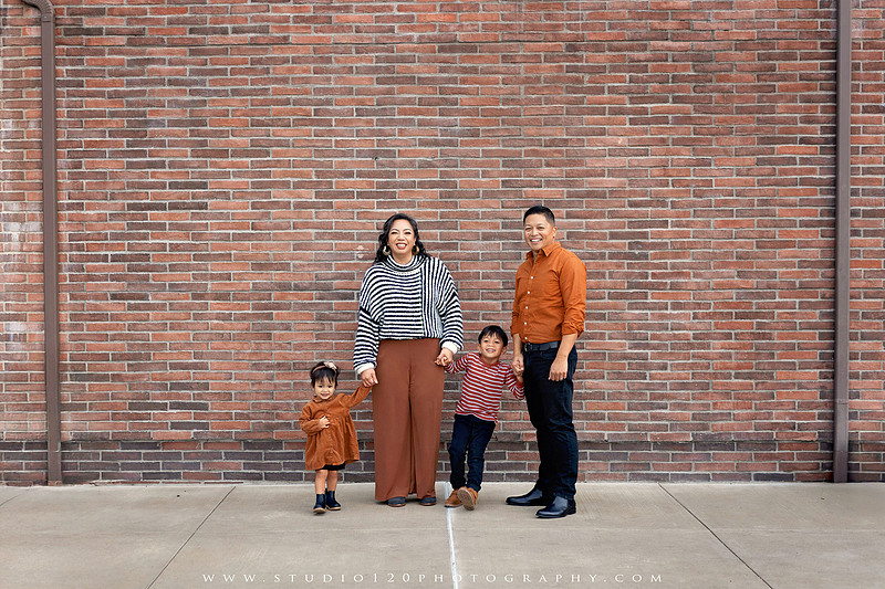 Outdoor family portrait session featuring parents and two young children smiling together against a warm neutral architectural backdrop