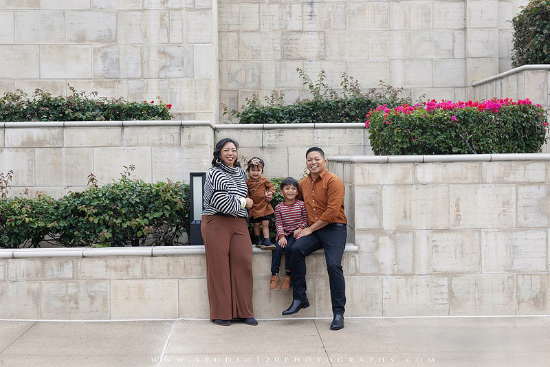 Outdoor family portrait session featuring parents and two young children smiling together against a warm neutral architectural backdrop