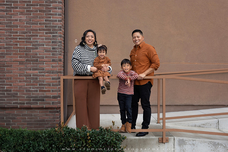 Outdoor family portrait session featuring parents and two young children smiling together against a warm neutral architectural backdrop