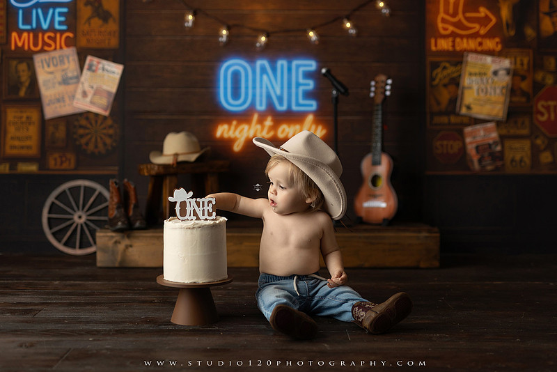 EvotoOne-year-old baby wearing a cowboy hat sits on a rustic honky-tonk stage with vintage posters, string lights, and a neon “One Night Only” sign during a country-themed milestone photo session.