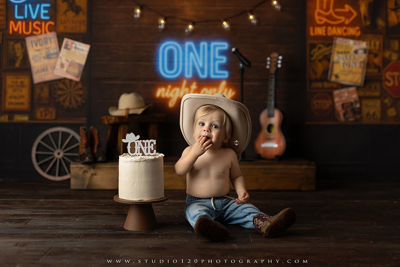 EvotoOne-year-old baby wearing a cowboy hat sits on a rustic honky-tonk stage with vintage posters, string lights, and a neon “One Night Only” sign during a country-themed milestone photo session.