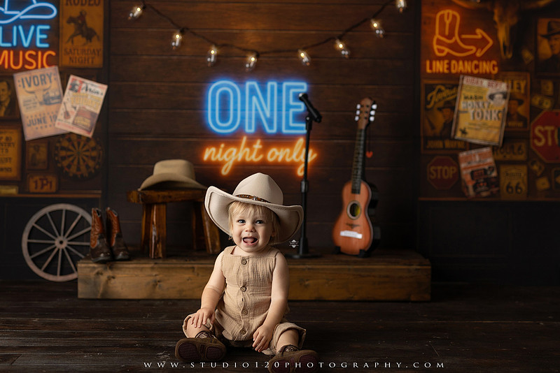 EvotoOne-year-old baby wearing a cowboy hat sits on a rustic honky-tonk stage with vintage posters, string lights, and a neon “One Night Only” sign during a country-themed milestone photo session.