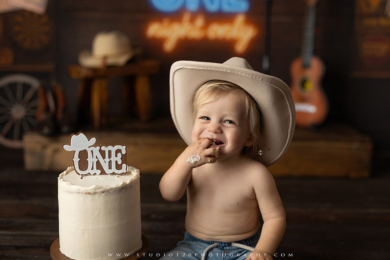 EvotoOne-year-old baby wearing a cowboy hat sits on a rustic honky-tonk stage with vintage posters, string lights, and a neon “One Night Only” sign during a country-themed milestone photo session.