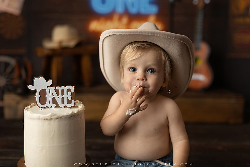 EvotoOne-year-old baby wearing a cowboy hat sits on a rustic honky-tonk stage with vintage posters, string lights, and a neon “One Night Only” sign during a country-themed milestone photo session.