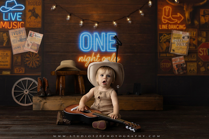 EvotoOne-year-old baby wearing a cowboy hat sits on a rustic honky-tonk stage with vintage posters, string lights, and a neon “One Night Only” sign during a country-themed milestone photo session.
