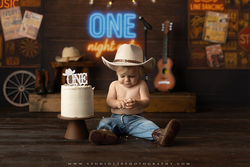 EvotoOne-year-old baby wearing a cowboy hat sits on a rustic honky-tonk stage with vintage posters, string lights, and a neon “One Night Only” sign during a country-themed milestone photo session.