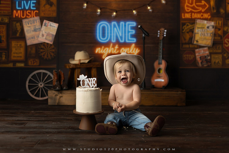EvotoOne-year-old baby wearing a cowboy hat sits on a rustic honky-tonk stage with vintage posters, string lights, and a neon “One Night Only” sign during a country-themed milestone photo session.