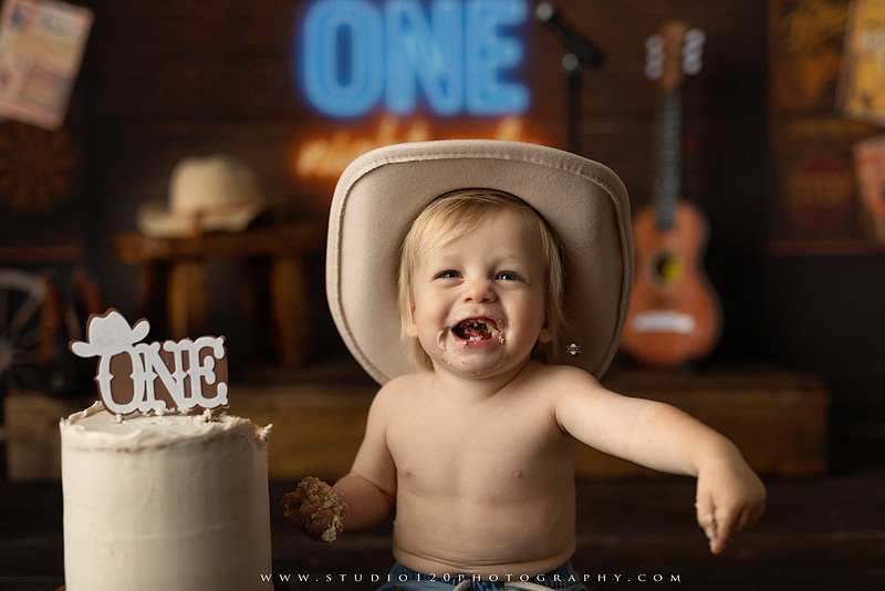 EvotoOne-year-old baby wearing a cowboy hat sits on a rustic honky-tonk stage with vintage posters, string lights, and a neon “One Night Only” sign during a country-themed milestone photo session.