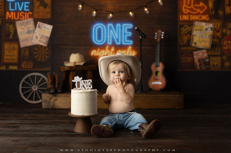 EvotoOne-year-old baby wearing a cowboy hat sits on a rustic honky-tonk stage with vintage posters, string lights, and a neon “One Night Only” sign during a country-themed milestone photo session.