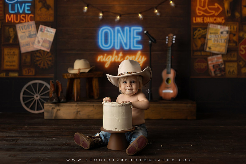 EvotoOne-year-old baby wearing a cowboy hat sits on a rustic honky-tonk stage with vintage posters, string lights, and a neon “One Night Only” sign during a country-themed milestone photo session.