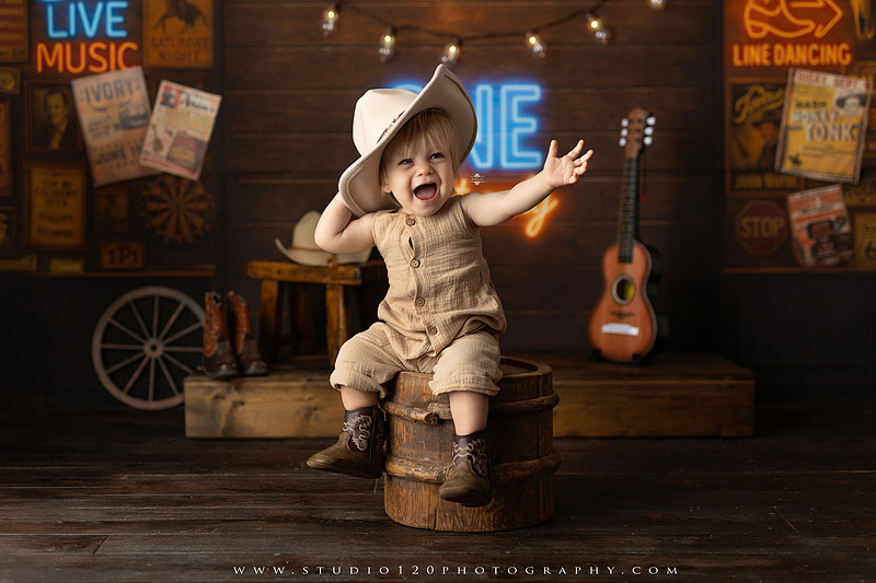 EvotoOne-year-old baby wearing a cowboy hat sits on a rustic honky-tonk stage with vintage posters, string lights, and a neon “One Night Only” sign during a country-themed milestone photo session.