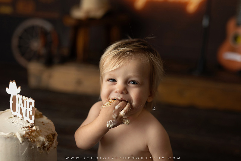 EvotoOne-year-old baby wearing a cowboy hat sits on a rustic honky-tonk stage with vintage posters, string lights, and a neon “One Night Only” sign during a country-themed milestone photo session.