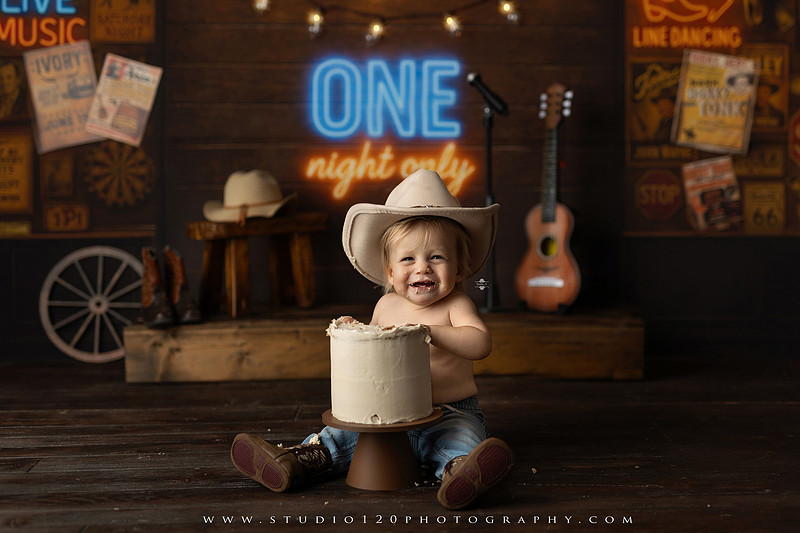 EvotoOne-year-old baby wearing a cowboy hat sits on a rustic honky-tonk stage with vintage posters, string lights, and a neon “One Night Only” sign during a country-themed milestone photo session.