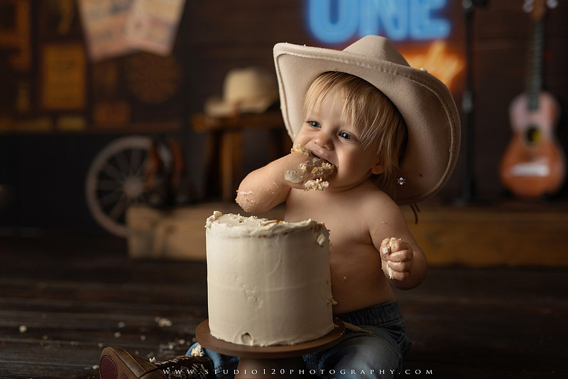 EvotoOne-year-old baby wearing a cowboy hat sits on a rustic honky-tonk stage with vintage posters, string lights, and a neon “One Night Only” sign during a country-themed milestone photo session.