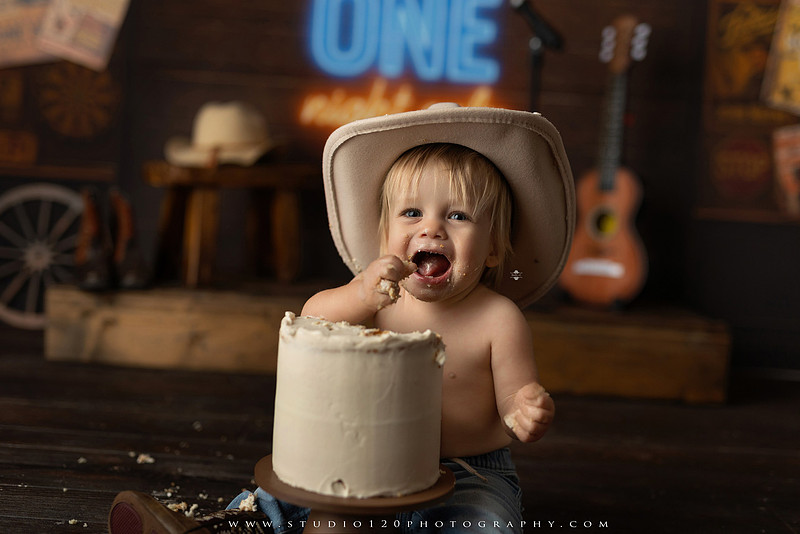 EvotoOne-year-old baby wearing a cowboy hat sits on a rustic honky-tonk stage with vintage posters, string lights, and a neon “One Night Only” sign during a country-themed milestone photo session.