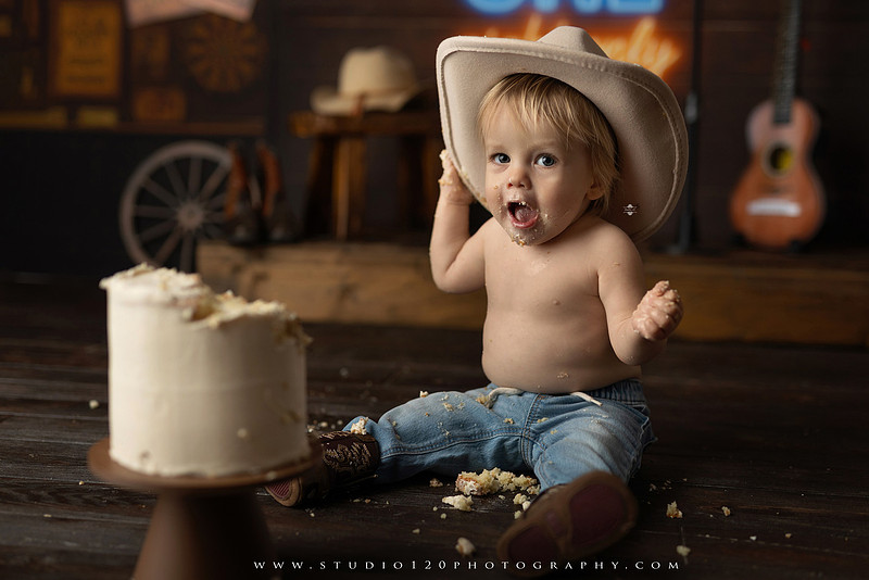 EvotoOne-year-old baby wearing a cowboy hat sits on a rustic honky-tonk stage with vintage posters, string lights, and a neon “One Night Only” sign during a country-themed milestone photo session.