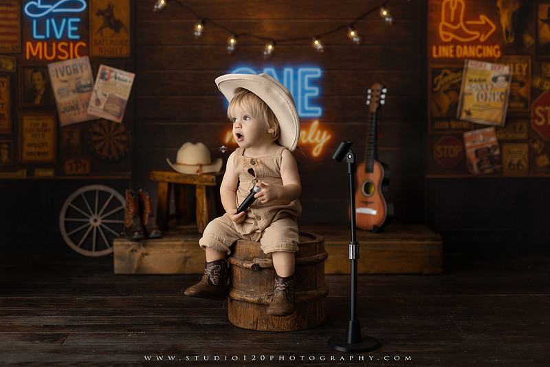 EvotoOne-year-old baby wearing a cowboy hat sits on a rustic honky-tonk stage with vintage posters, string lights, and a neon “One Night Only” sign during a country-themed milestone photo session.