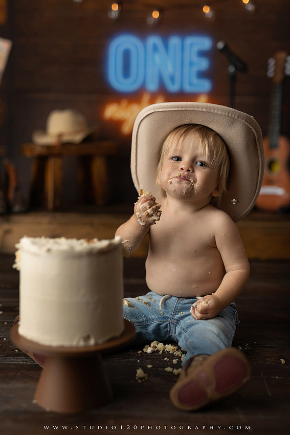 EvotoOne-year-old baby wearing a cowboy hat sits on a rustic honky-tonk stage with vintage posters, string lights, and a neon “One Night Only” sign during a country-themed milestone photo session.