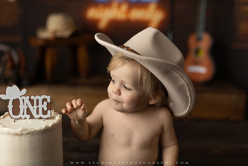 EvotoOne-year-old baby wearing a cowboy hat sits on a rustic honky-tonk stage with vintage posters, string lights, and a neon “One Night Only” sign during a country-themed milestone photo session.