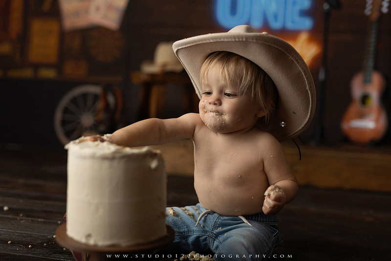EvotoOne-year-old baby wearing a cowboy hat sits on a rustic honky-tonk stage with vintage posters, string lights, and a neon “One Night Only” sign during a country-themed milestone photo session.