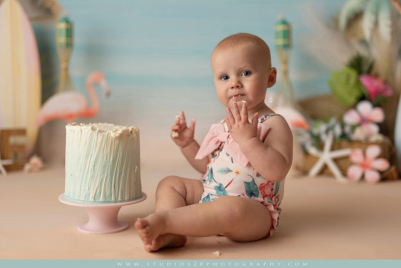 “first birthday portrait of baby in white dress with floral accents in light and airy studio”
“elegant first birthday portrait session with baby in white dress and neutral backdrop”
“luxe coastal cake smash with beach themed backdrop, flamingos and tropical decor”
“baby girl beach themed cake smash with pastel cake and tropical surfboard props”
“coastal first birthday cake smash with flamingo accents and soft beach colors”
“tropical cake smash photography featuring baby and pastel beach themed set”
“baby enjoying cake smash in coastal themed studio with palm trees and flamingos”
“beach inspired cake smash session with baby standing in cake and tropical decor”
“luxury cake smash photography with coastal beach theme and soft pastel tones”