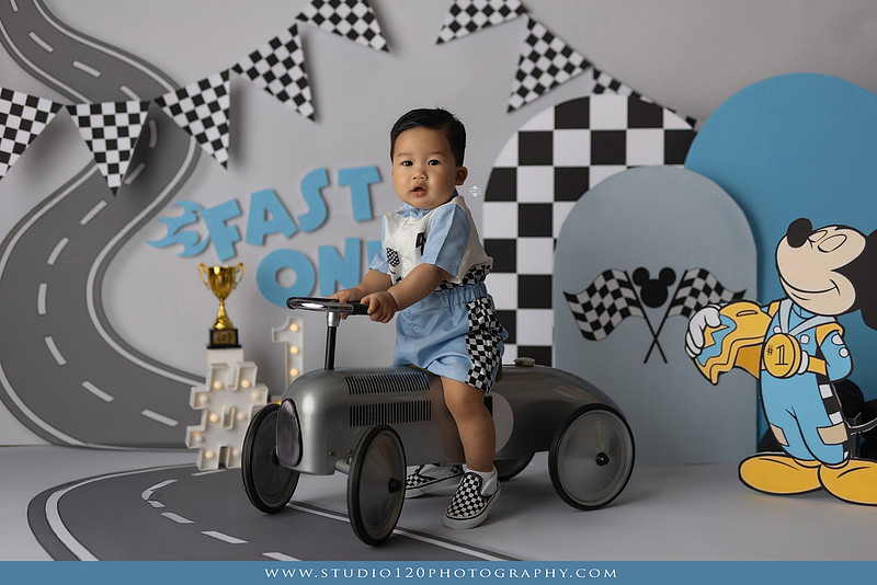 Fast One Mickey Mouse race car cake smash featuring custom race track backdrop, checkered flags, vintage race car prop, and first birthday cake in San Diego