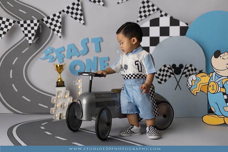 Fast One Mickey Mouse race car cake smash featuring custom race track backdrop, checkered flags, vintage race car prop, and first birthday cake in San Diego
