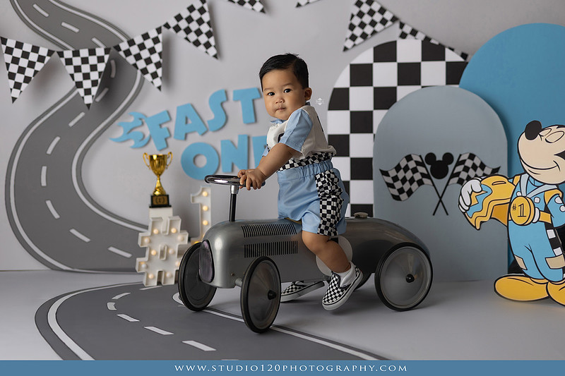 Fast One Mickey Mouse race car cake smash featuring custom race track backdrop, checkered flags, vintage race car prop, and first birthday cake in San Diego