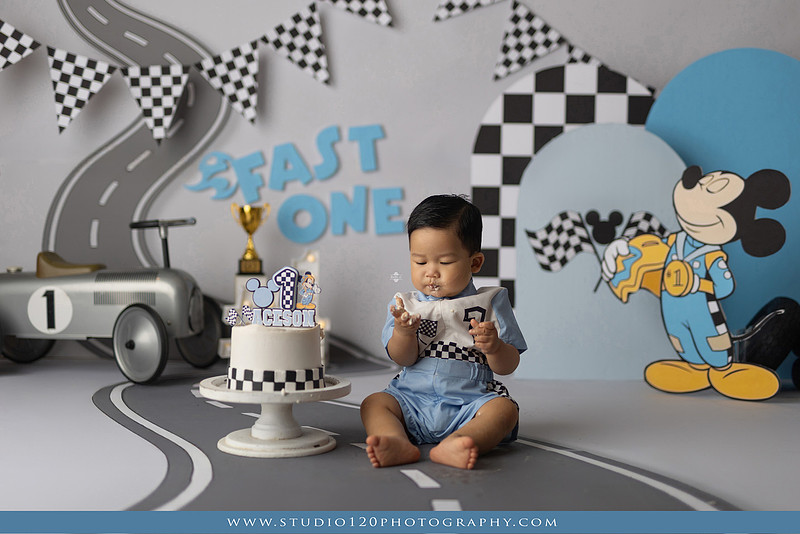 Fast One Mickey Mouse race car cake smash featuring custom race track backdrop, checkered flags, vintage race car prop, and first birthday cake in San Diego