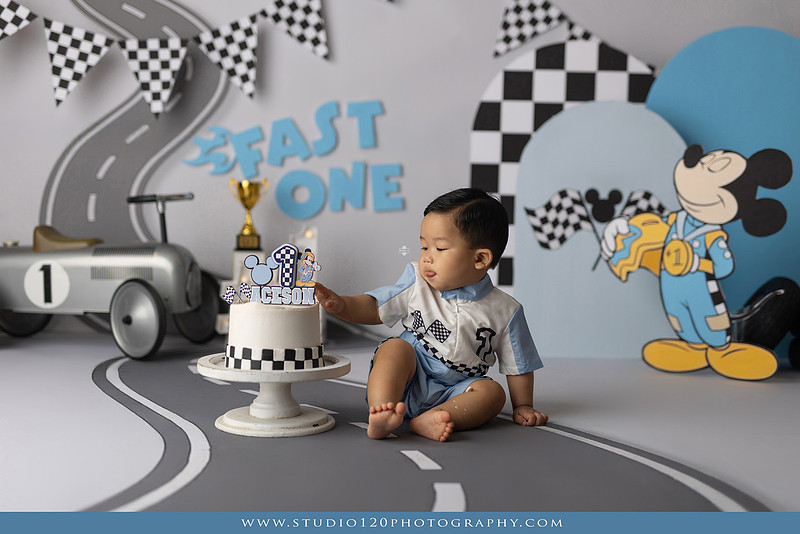 Fast One Mickey Mouse race car cake smash featuring custom race track backdrop, checkered flags, vintage race car prop, and first birthday cake in San Diego