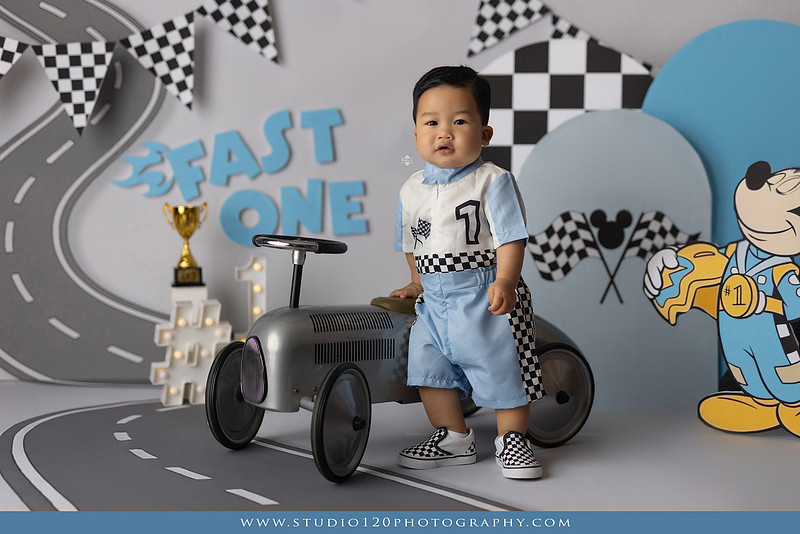 Fast One Mickey Mouse race car cake smash featuring custom race track backdrop, checkered flags, vintage race car prop, and first birthday cake in San Diego