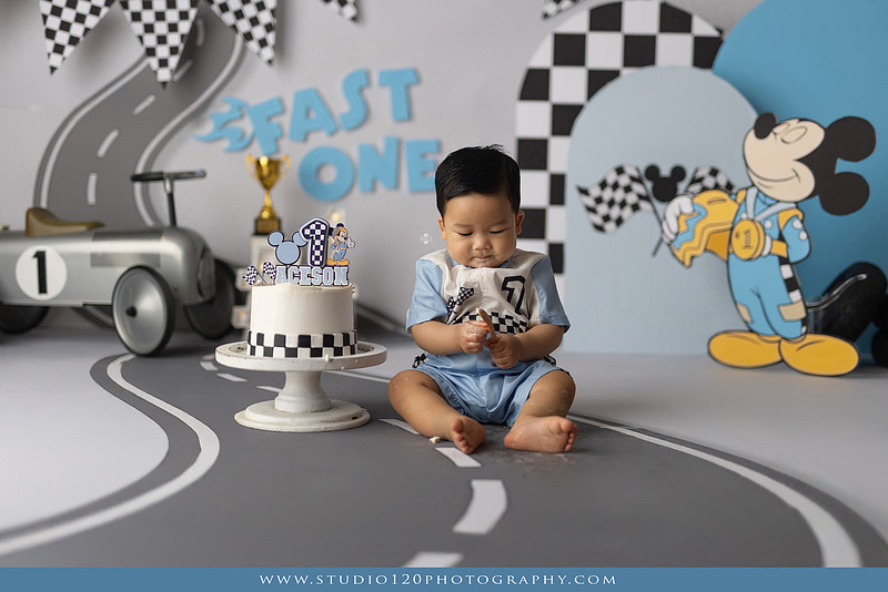 Fast One Mickey Mouse race car cake smash featuring custom race track backdrop, checkered flags, vintage race car prop, and first birthday cake in San Diego
