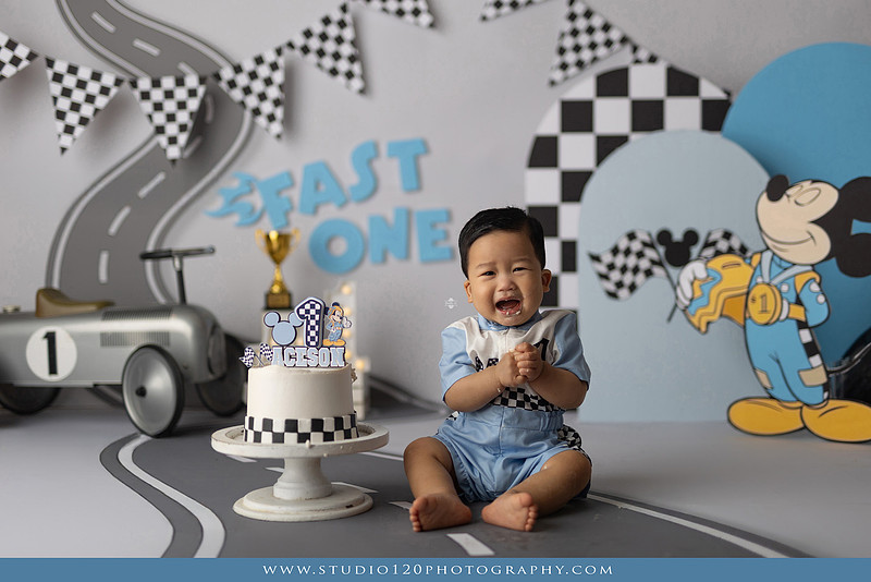 Fast One Mickey Mouse race car cake smash featuring custom race track backdrop, checkered flags, vintage race car prop, and first birthday cake in San Diego