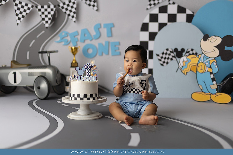 Fast One Mickey Mouse race car cake smash featuring custom race track backdrop, checkered flags, vintage race car prop, and first birthday cake in San Diego